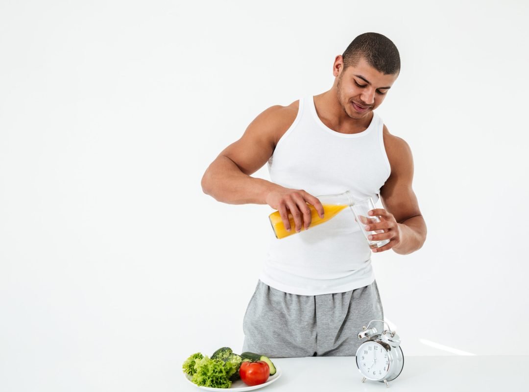Image of handsome young sportsman standing over white background holding glass of juice. Looking aside.