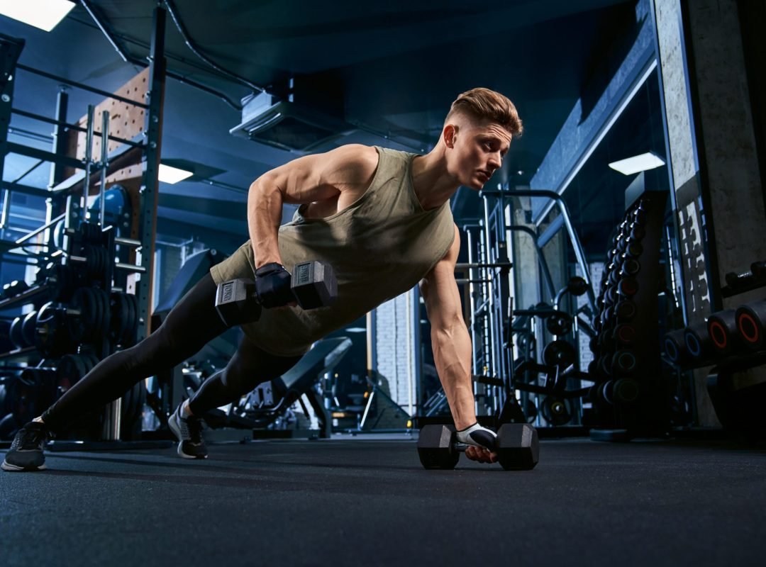 Muscular caucasian man training on floor using dumbbells in sports club. Close up of young sportsman building muscles, doing push ups on one hand in empty gym. Concept of bodybuilding.
